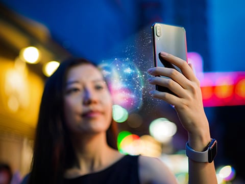 An out-of-focus woman on a city street at night faces her mobile device while a seemingly holographic grid maps her features between her face and a mobile phone in her hand